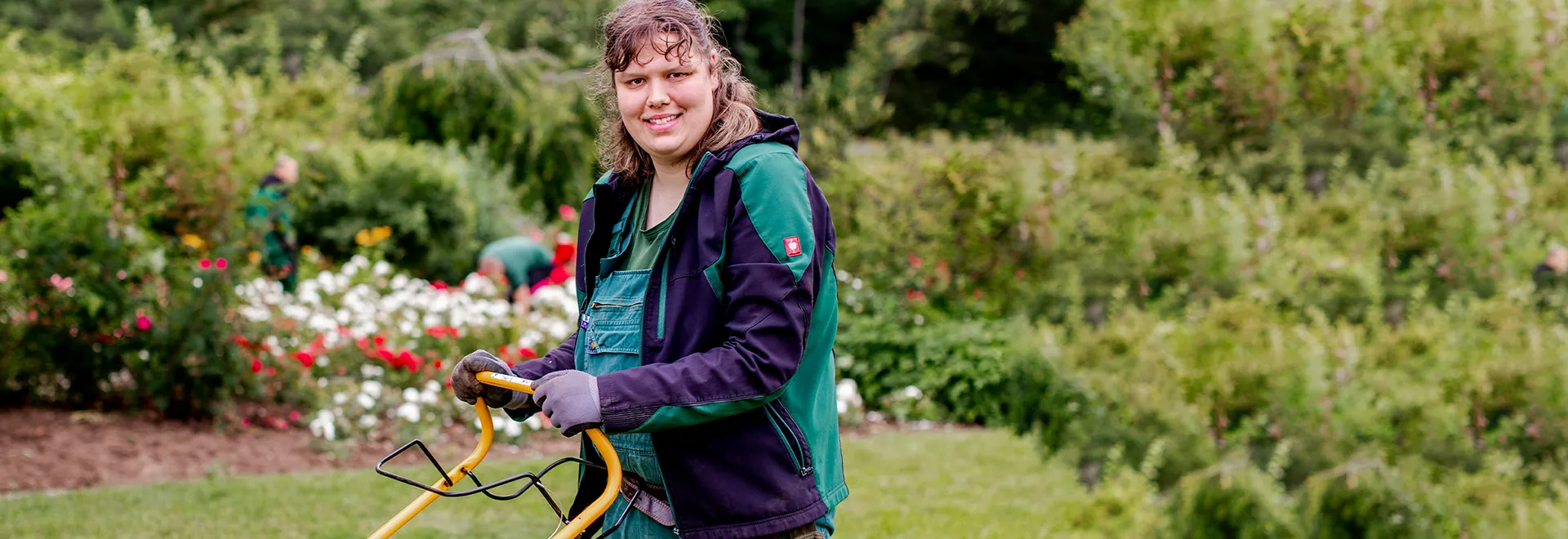 Eine Frau in Arbeitskleidung mäht mit einem Rasenmäher einen Garten.