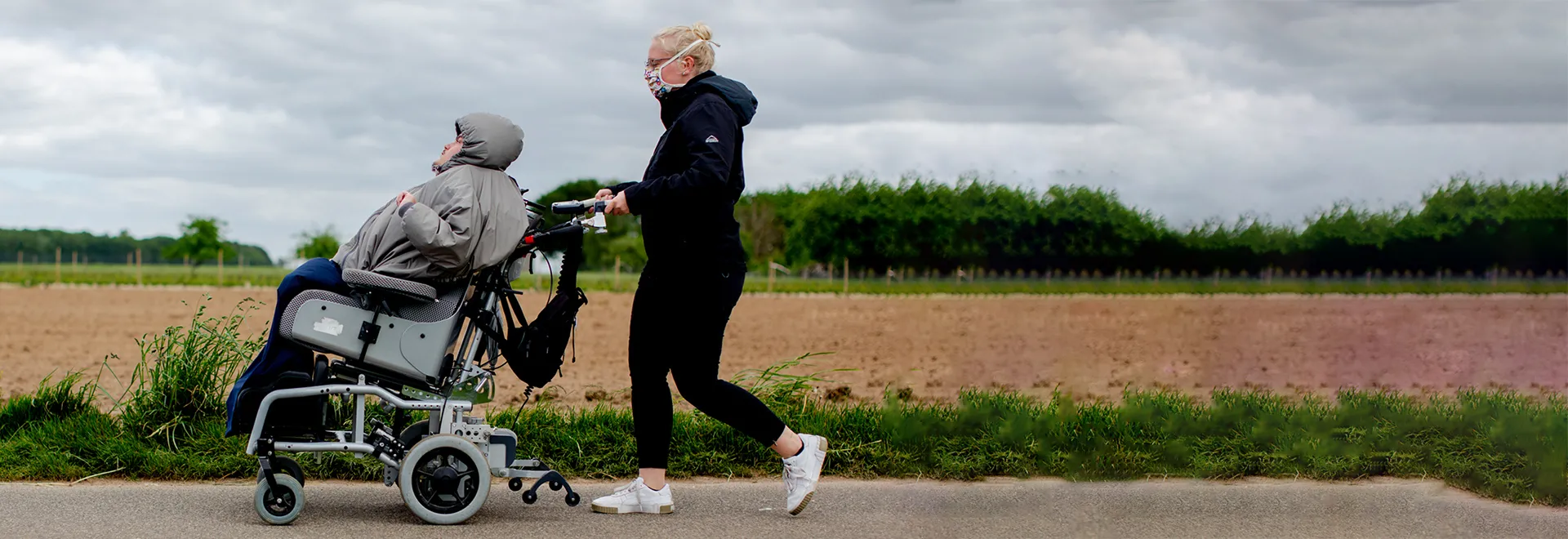 Eine Frau mit Mundschutz schiebt einen Mann im Rollstuhl an einem Feld vorbei.