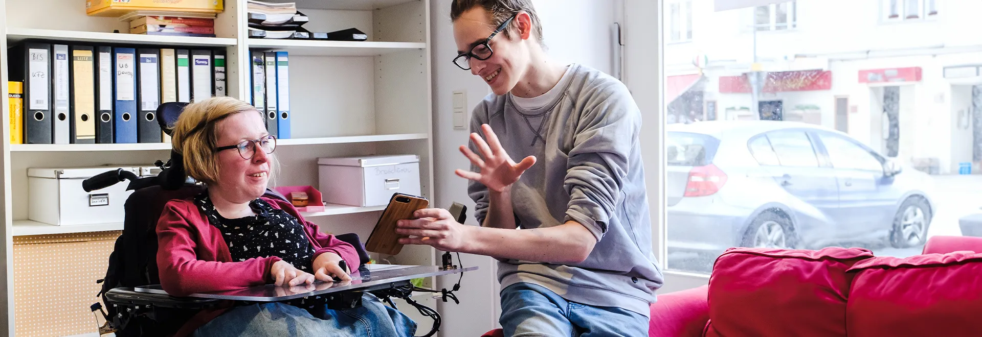 Ein junger Mann zeigt einer Frau im Rollstuhl etwas auf seinem Handy.
