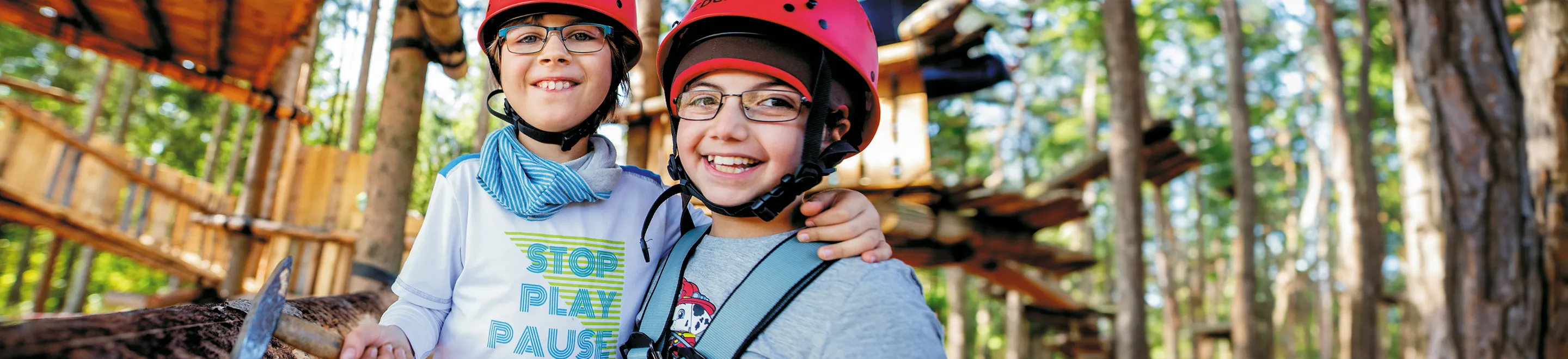 Zwei Kinder stehen in einem Abenteuerspielplatz im Wald, haben einen Helm auf dem Kopf, eines einen Hammer in der Hand.