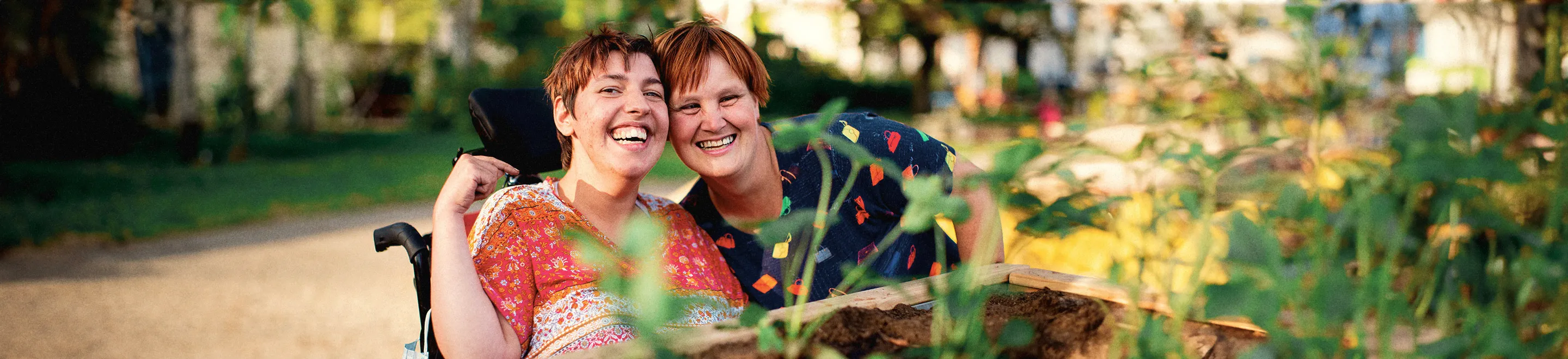 Zwei Frauen in bunt gemusterten Oberteilen sind hinter einem Hochbeet zu sehen. Eine der beiden Frauen sitzt in einem Rollstuhl. Sie stecken die Köpfe zusammen und lachen in die Kamera.