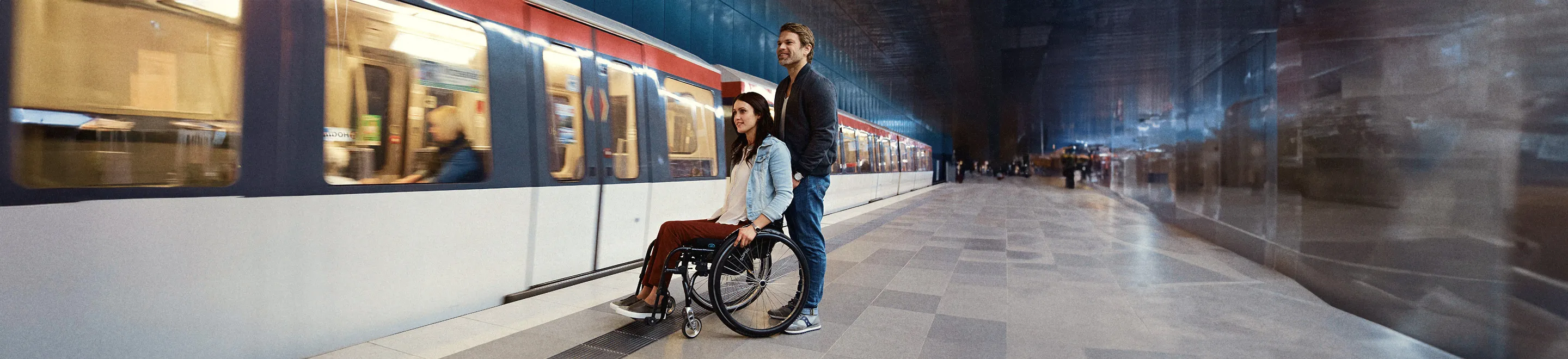 Eine Frau im Rollstuhl und ein Mann hinter ihr stehen vor einer einfahrenden U-Bahn.