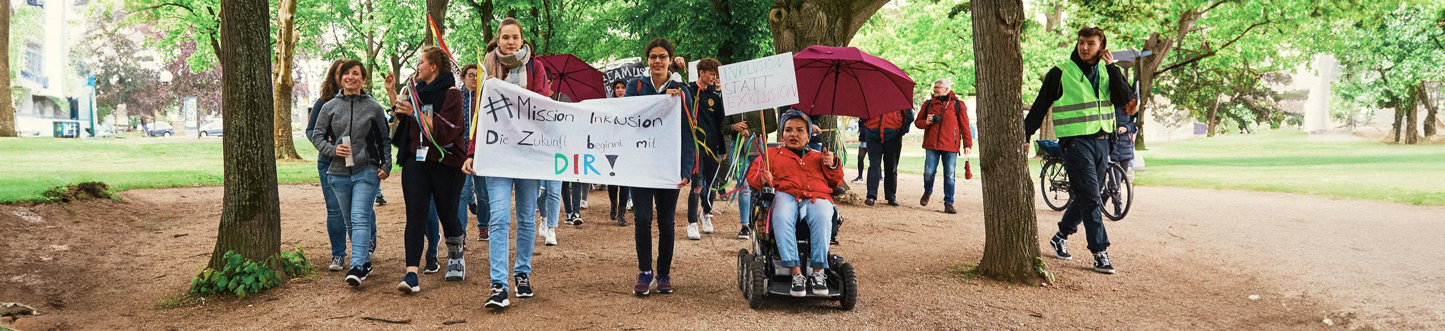 Eine Gruppe junger Demonstranten, die durch einen Park laufen. Sie halten ein Transparent auf dem steht: "#Mission Inklusion - Die Zukunft beginnt bei Dir".