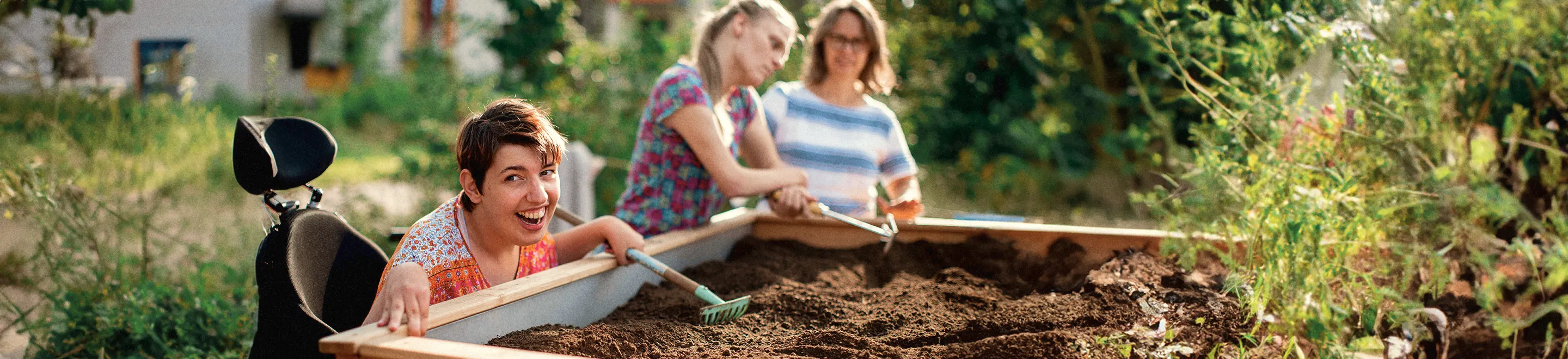 Links im Vordergrund ist eine lachende Frau im Rollstuhl, die mit einer Harke die Erde eines Hochbeets harkt. Im Hintergrund stehen zwei weitere Frauen am Hochbeet.