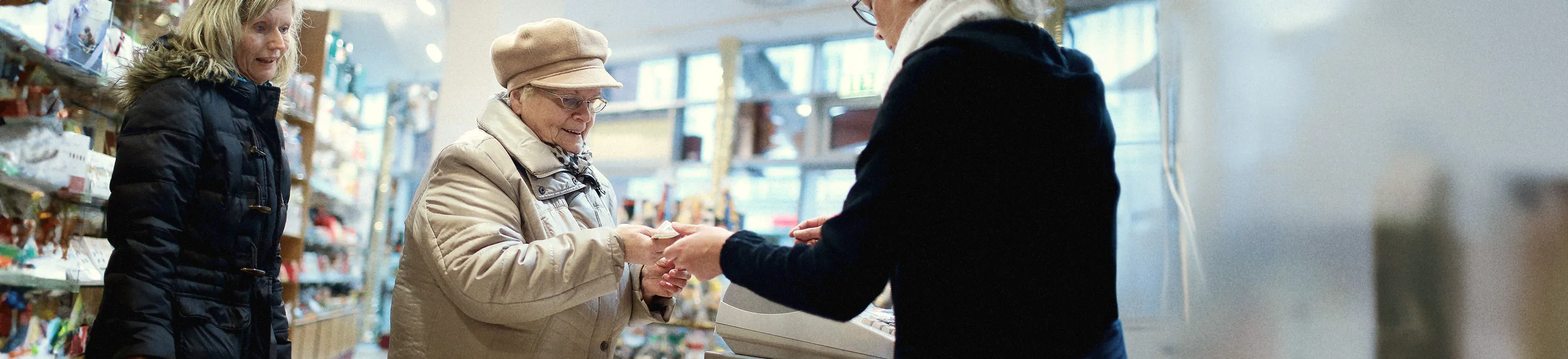 Eine ältere Frau mit beigem Mantel und Hut nimmt an einer Kasse Rückgeld entgegen, das ihr eine Kassiererin reicht.