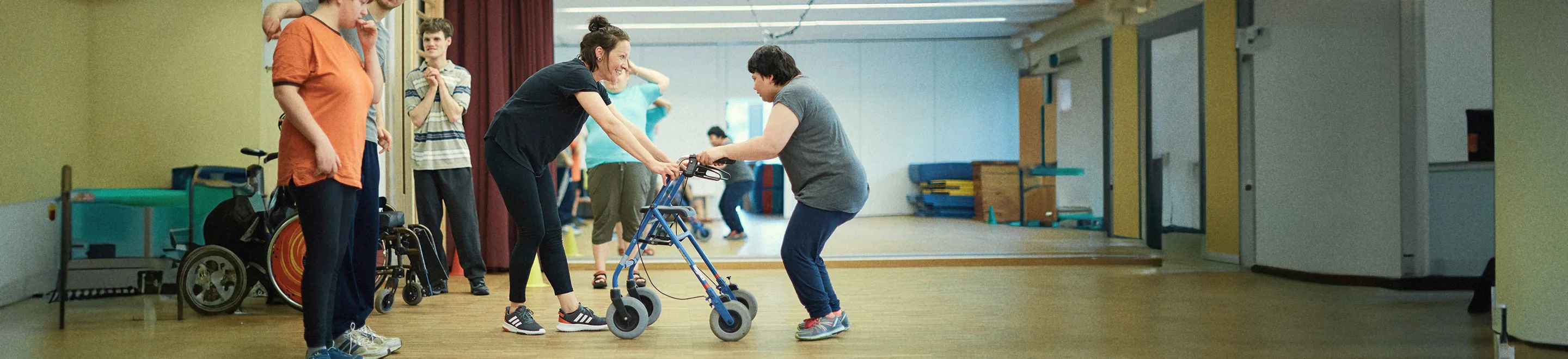 Eine Frau übt an einem Rollator in einem Gymnastikraum zu gehen. Eine Trainerin hält den Rollator für sie fest und lächelt sie an.