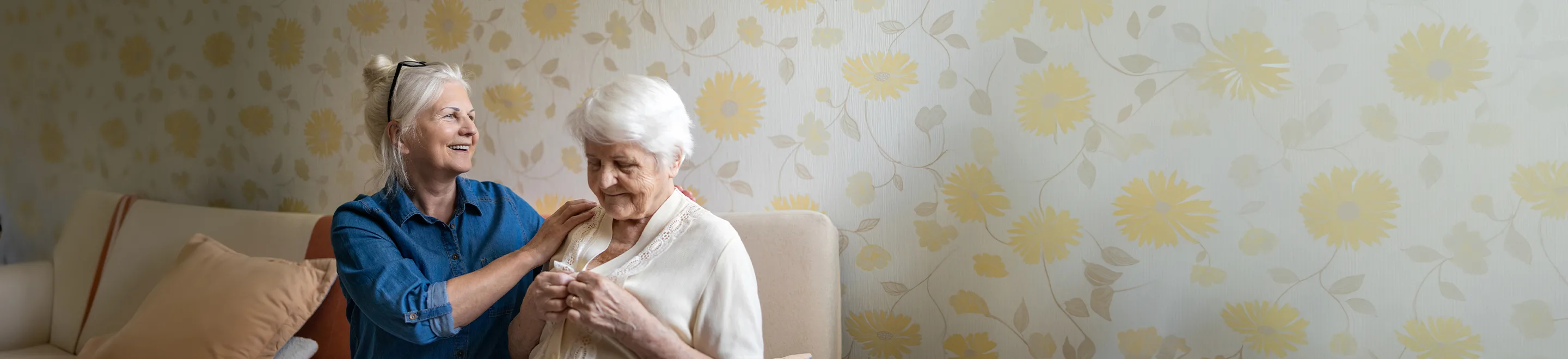 Eine Frau hilft einer Seniorin in einem Wohnzimmer beim Ankleiden.