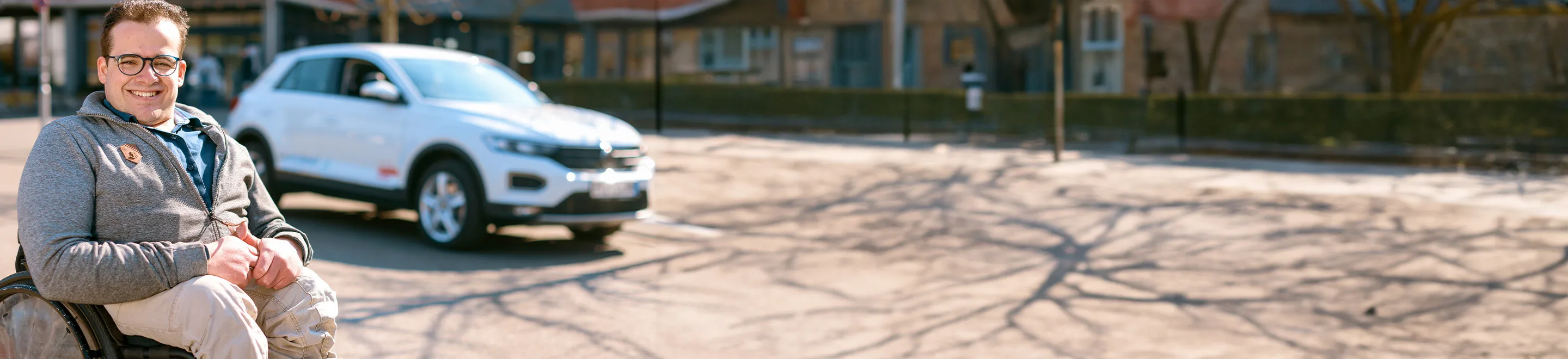 Ein Mann sitzt im Rollstuhl und lächelt. Im Hintergrund steht ein Auto auf einem Parkplatz.