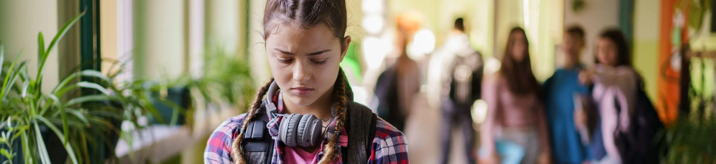 Ein Mädchen geht mit gesenktem Kopf und Blick durch den Gang einer Schule. Im Hintergrund stehen drei weitere Schülerinnen. Eine von ihnen zeigt mit dem Finger auf das Mädchen.