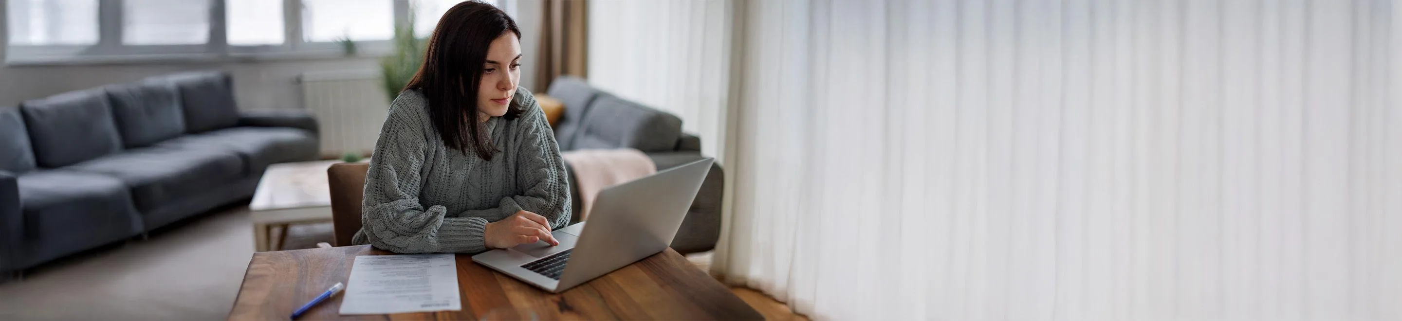 Eine Frau sitzt an einem Tisch und arbeitet an einem Laptop. Neben ihr sind ein Formular und ein Stift zu sehen.