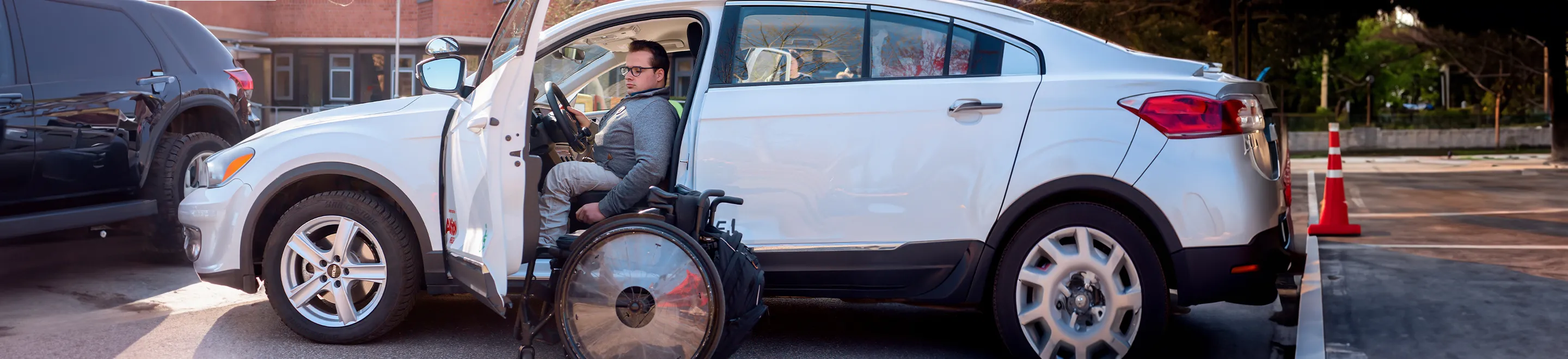 Ein junger Mann mit einer Gehbehinderung sitzt in einem Auto. Neben der geöffneten Fahrertür steht ein Rollstuhl.