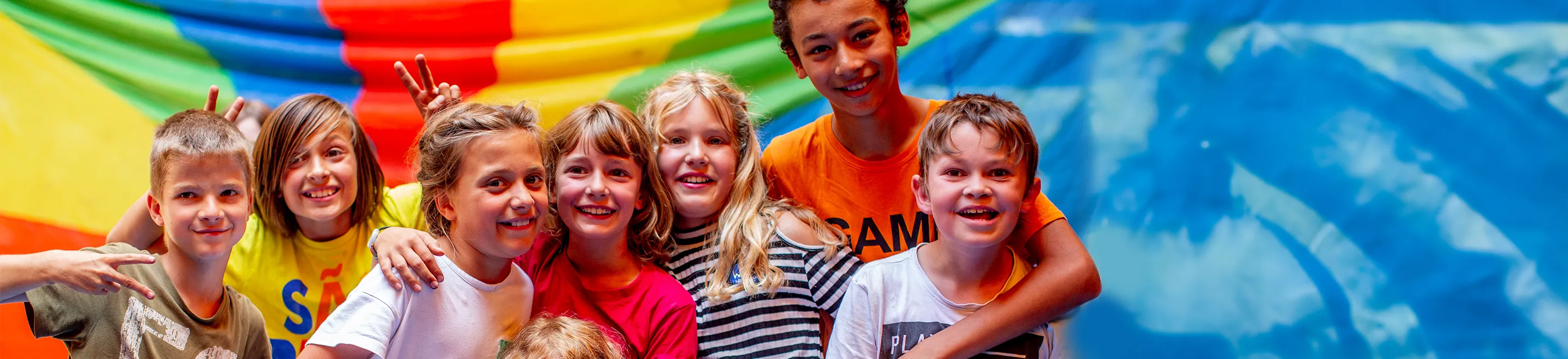 Mehrere fröhliche Kinder stehen vor einer bunten Fahne.