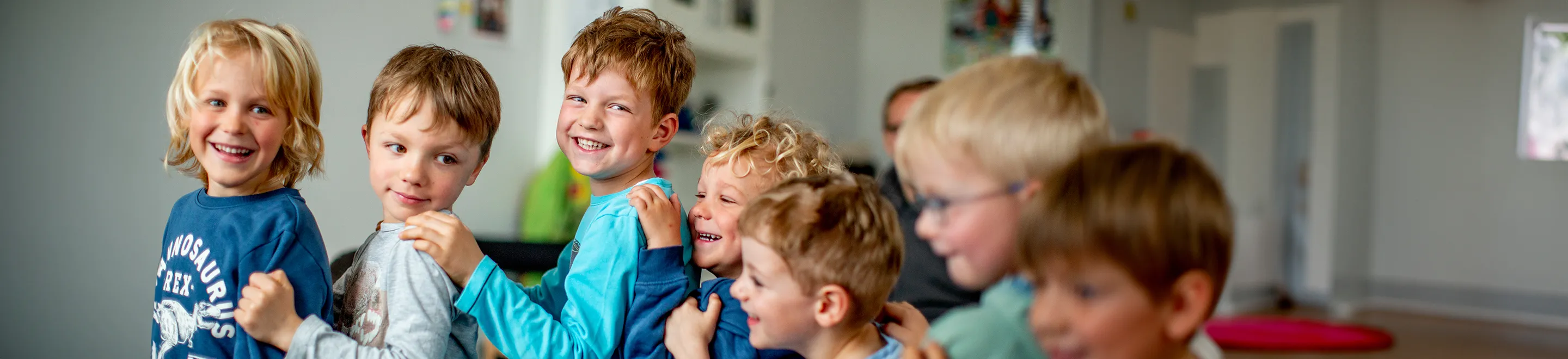 Mehrere fröhliche Kinder stehen hintereinander und fassen sich jeweils von hinten an die Schultern.