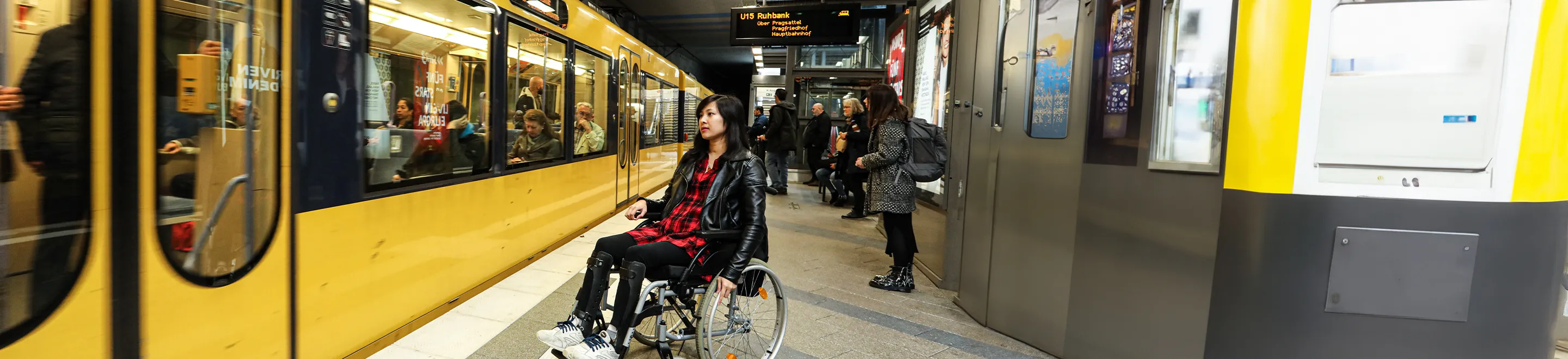 Eine junge Frau im Rollstuhl auf einem Bahnsteig. Eine U-Bahn fährt ein.