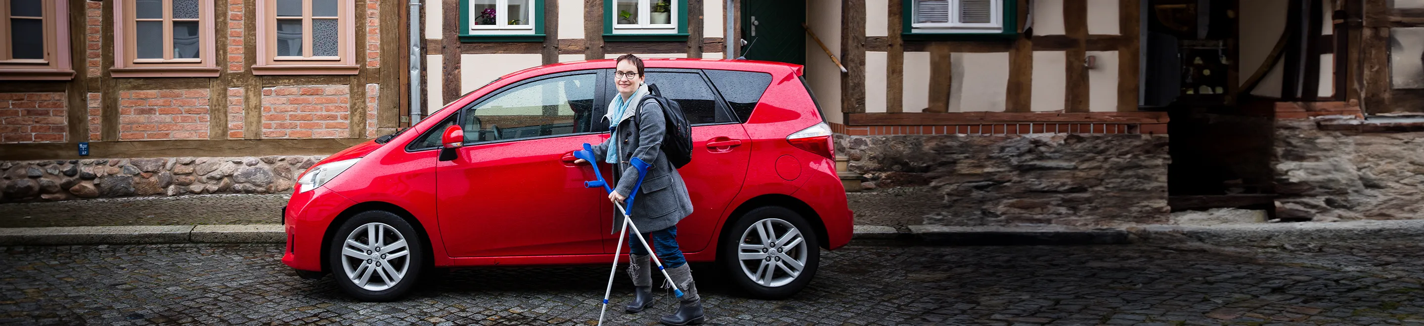 Eine Frau mit einer Gehbehinderung steht an der Fahrertür eines roten PKWs.
