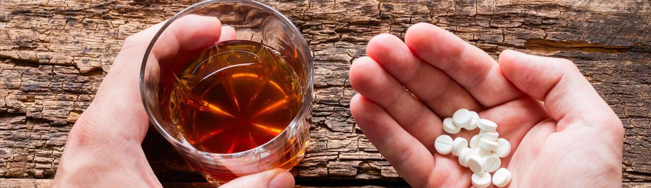 Zwei Hände; in der einen Hand liegen Tabletten, die andere Hand hält ein Glas mit Alkohol.