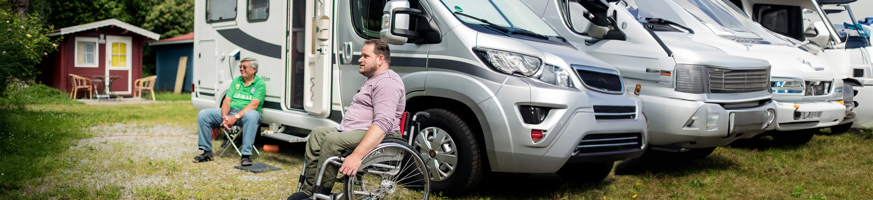 Zwei Männer sitzen vor einem Wohnmobil, das auf einem Campingplatz steht. Einer der beiden sitzt in einem Rollstuhl.