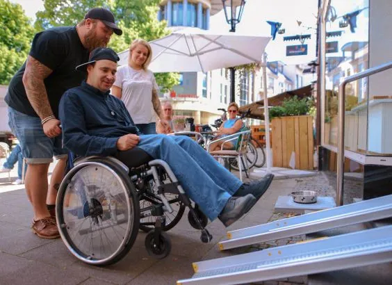 Ein Assistent schiebt eine Person im Rollstuhl eine Rampe hoch.