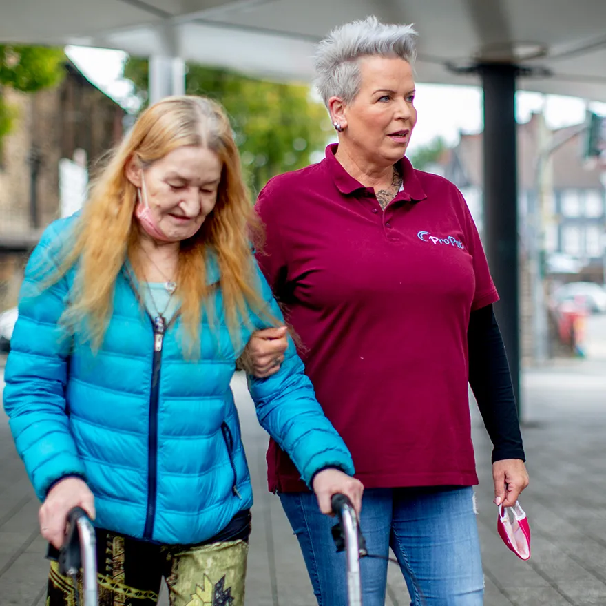 Eine Mitarbeiterin des Familienunterstützenden Dienstes stützt eine ältere Frau, die mit einem Rollator geht.