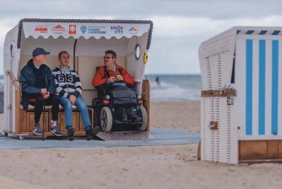 Drei Menschen sitzen in einem Strandkorb am Strand von Rostock, ein Mann sitzt im Rollstuhl.