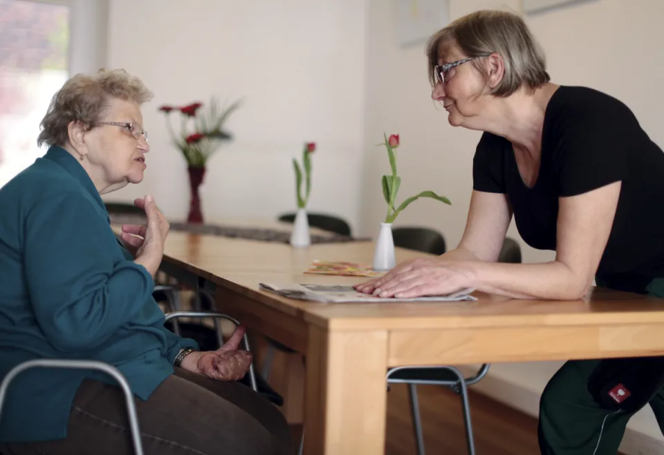 Eine Seniorin sitzt an einem Tisch. Ihr gegenüber steht eine Frau. Beide sprechen miteinander.
