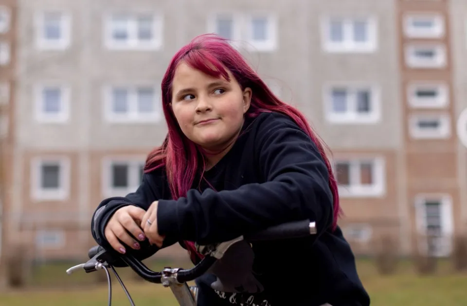 Eine Teenagerin steht mit einem Fahrrad vor einem Häuserblock und schaut skeptisch nach rechts.