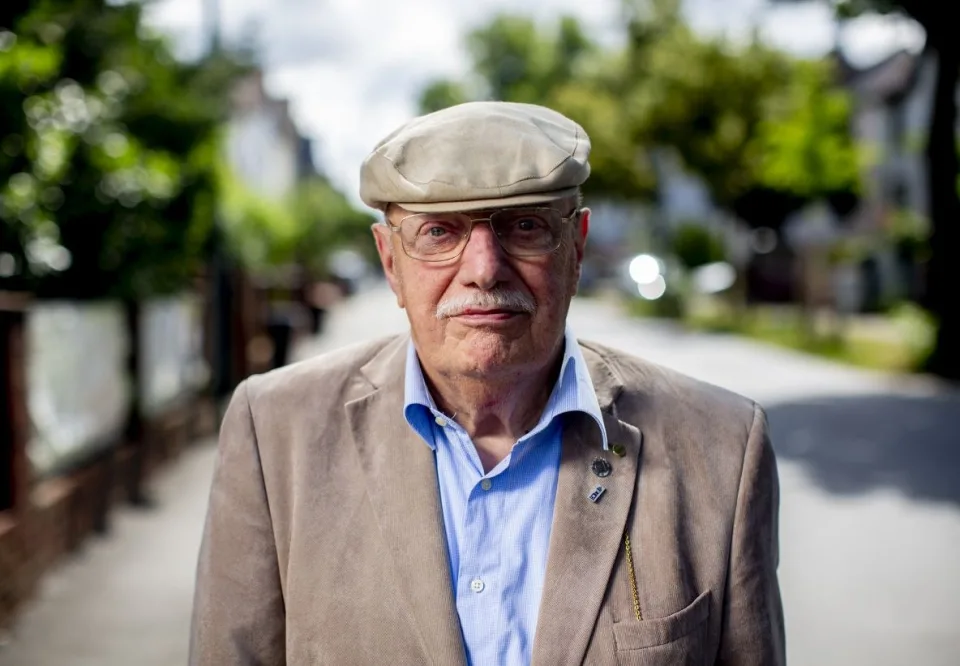 Ein älterer Mann mit Hut, grauen Haaren und Brille schaut ernst in die Kamera.