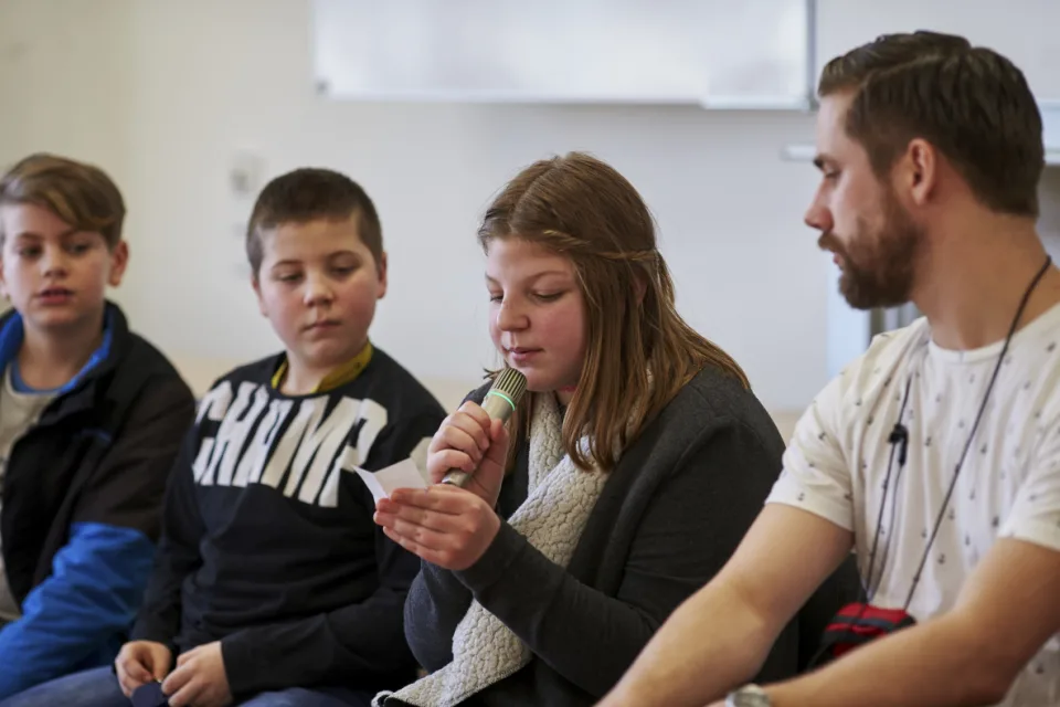 Mehrere Kinder und ein erwachsener Mann in einer Schulklasse. Ein Mädchen liest etwas vor und spricht in ein Mikrofon.