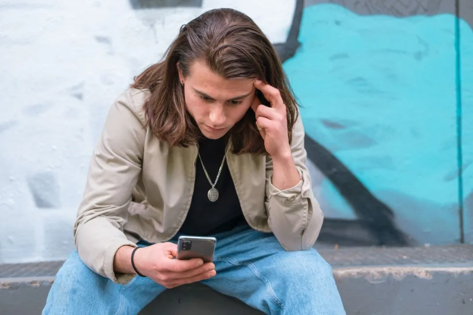 Ein junger Mann mit langen Haaren sitzt auf einer Stufe und schaut ernst in sein Handy.