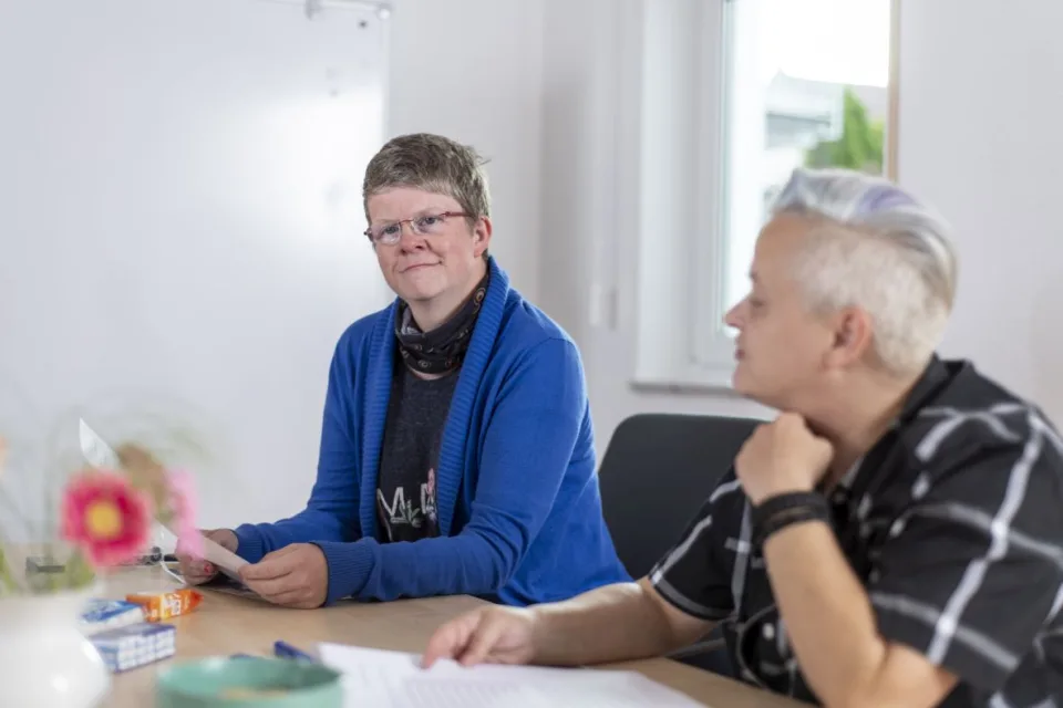 Zwei Frauen sitzen in einem Raum und halten ausgedruckte Papiere in der Hand.