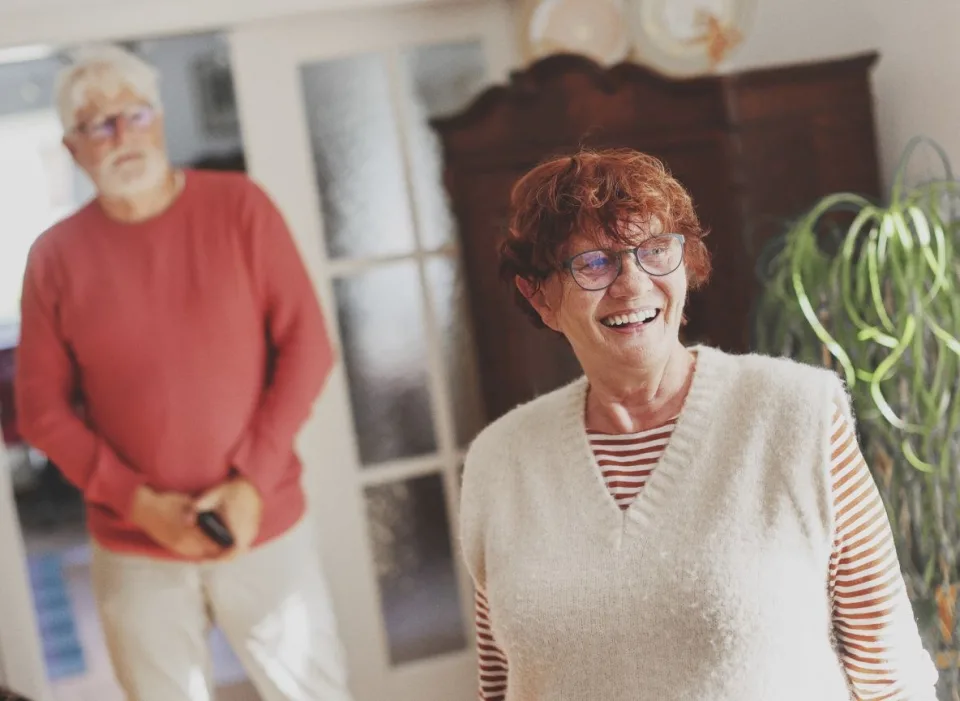Eine ältere Damen steht in einem Wohnzimmer und lächelt. Im Hintergrund steht ein älterer Mann.