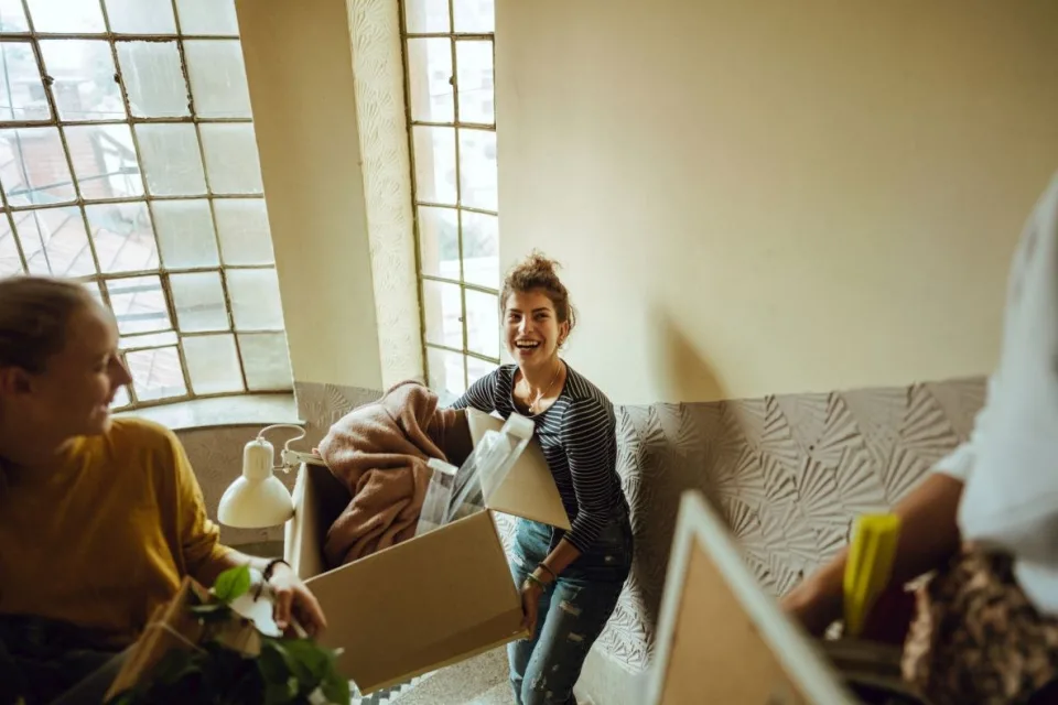 Eine junge Frau steht mit Umzugskisten im Treppenhaus eines Mehrfamilienhauses. Neben ihr sind weitere Personen, die ebenfalls Kisten in der Hand tragen. Alle lachen fröhlich.