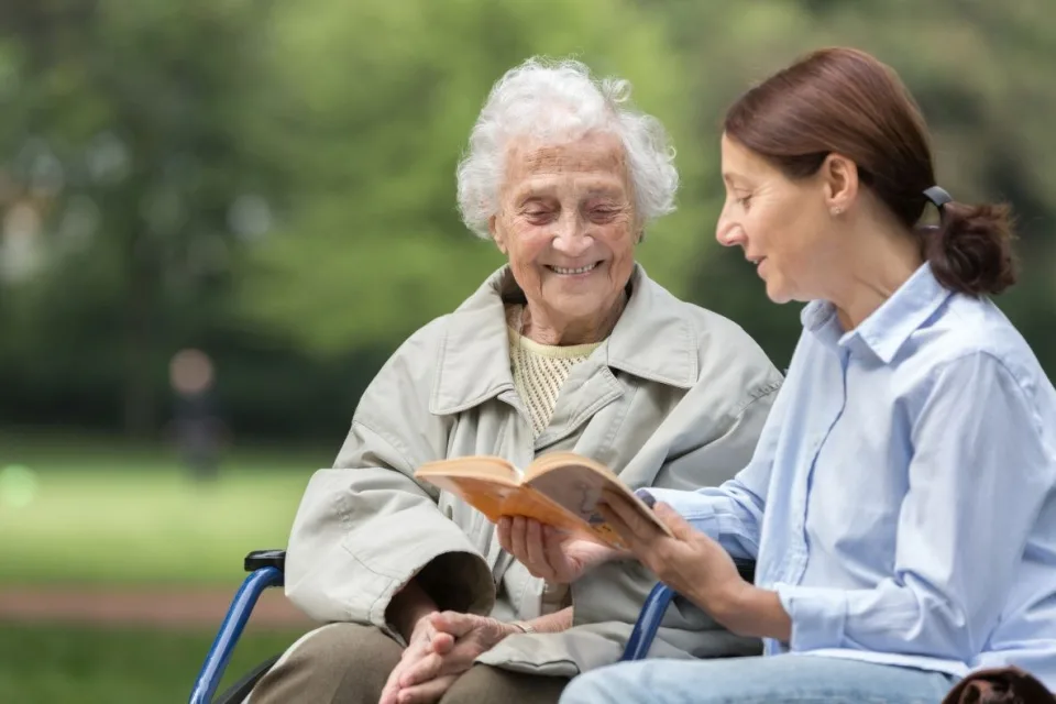 Eine Frau mittleren Alters und eine alte Frau sitzen in einem Park. Die ältere Frau im Rollstuhl. Die Frau mittleren Alters liest aus einem Buch vor.