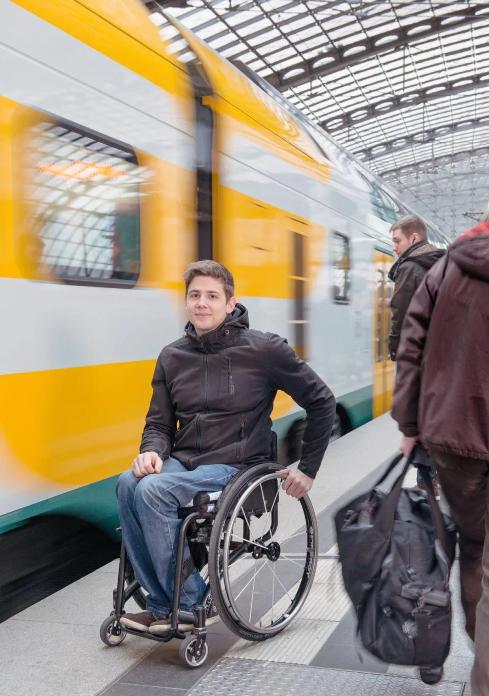 Ein junger Mann im Rollstuhl steht auf einem Bahnsteig. Daneben ein Zug.