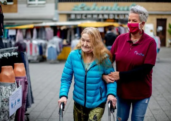 Eine Frau begleitet eine ältere Frau, die mit einer Gehhilfe geht, beim Einkaufen.