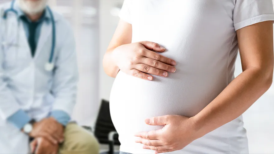Eine schwangere Frau hält ihren Bauch mit beiden Händen, im Hintergrund sitzt ein Arzt.