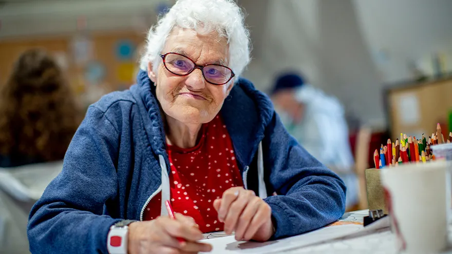Eine ältere Frau malt mit Bundstiften auf ein Blatt Papier.
