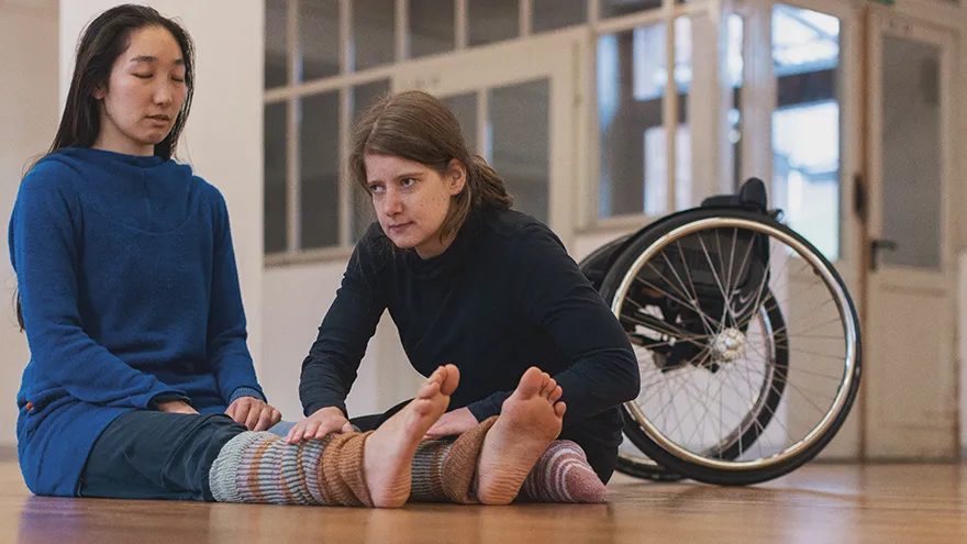 Zwei Frauen sitzen auf dem Fußboden, daneben steht ein Rollstuhl.