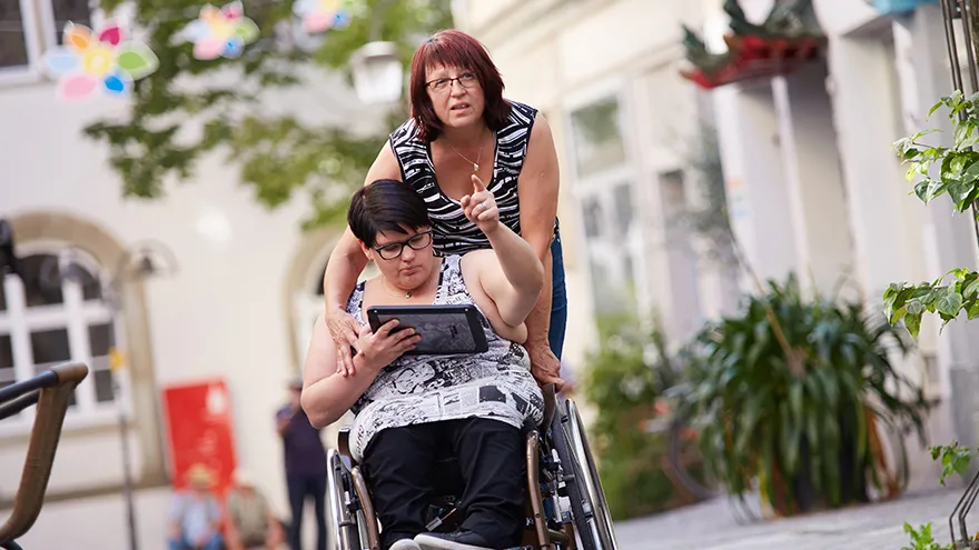 Eine Frau im Rollstuhl hat einen Tablet-PC in der Hand und weist mit der anderen Hand den Weg. Dahinter eine andere Frau, die den Rollstuhl schiebt.