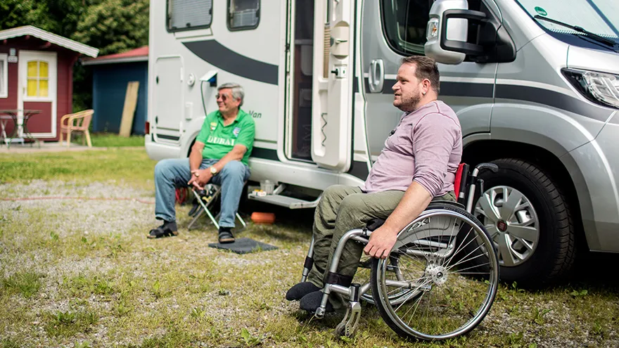 Ein Mann im Rollstuhl und ein Mann auf einem Hocker vor einem Wohnmobil, auf einem Campingplatz.