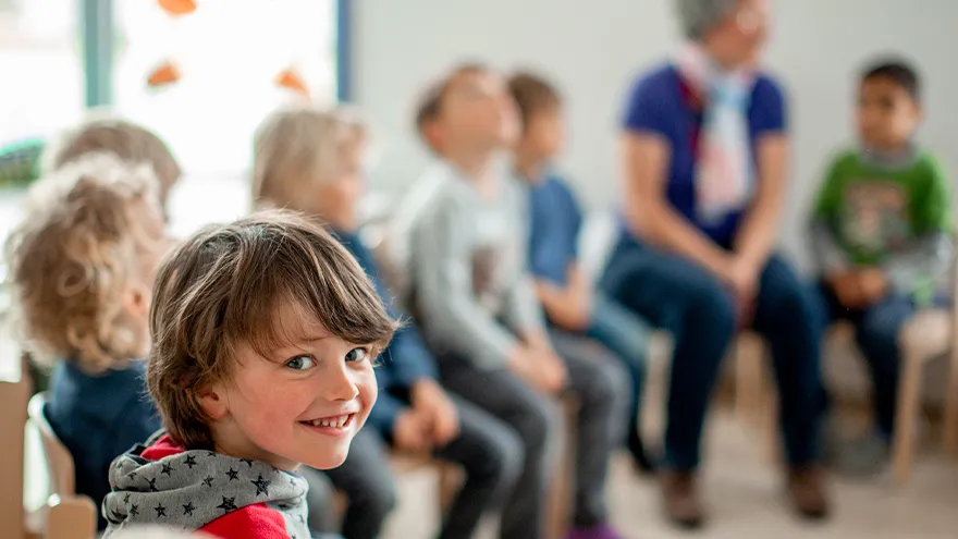 Ein Kind lächelt in die Kamera, es sitzt mit anderen Kindern und einer Frau in einem Stuhl-halbkreis.