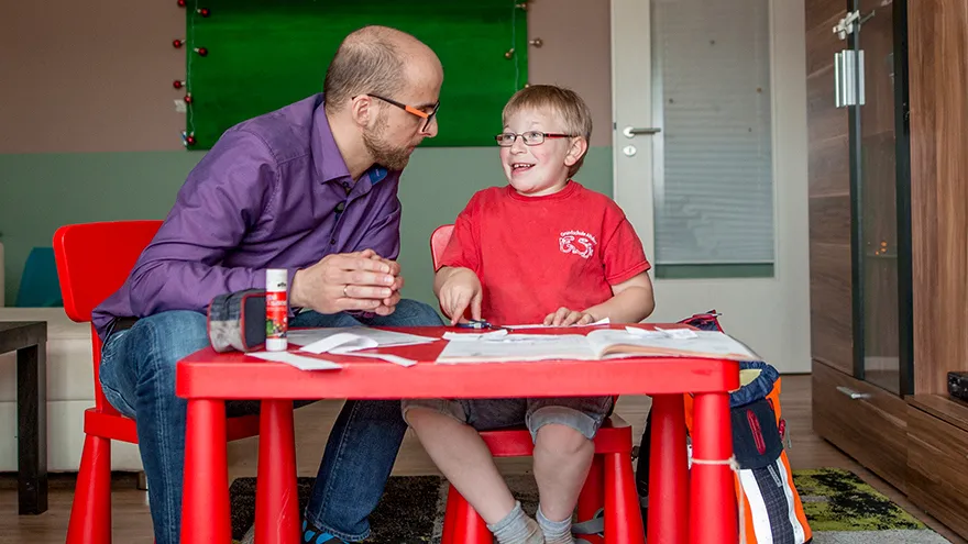 Ein Junge sitzt mit einem Mann an einem roten Tisch, sie unterhalten sich.