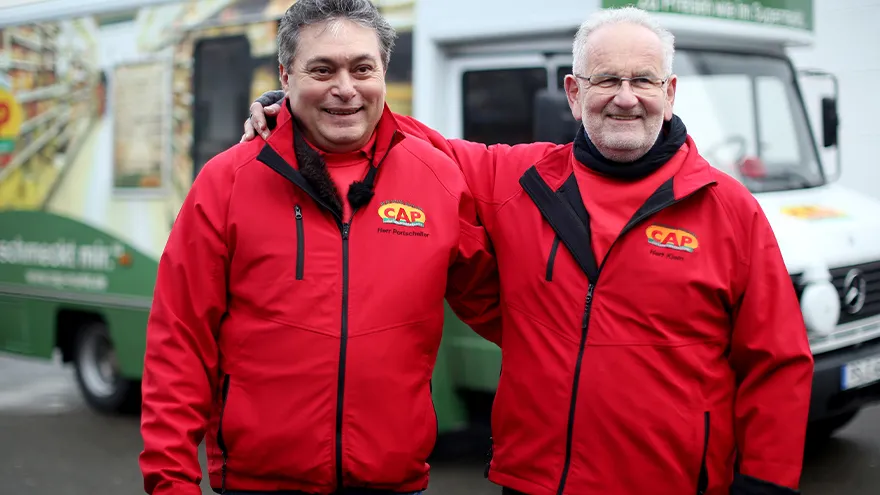 Zwei Männer in roten Jacken stehen vor einem Lieferwagen.