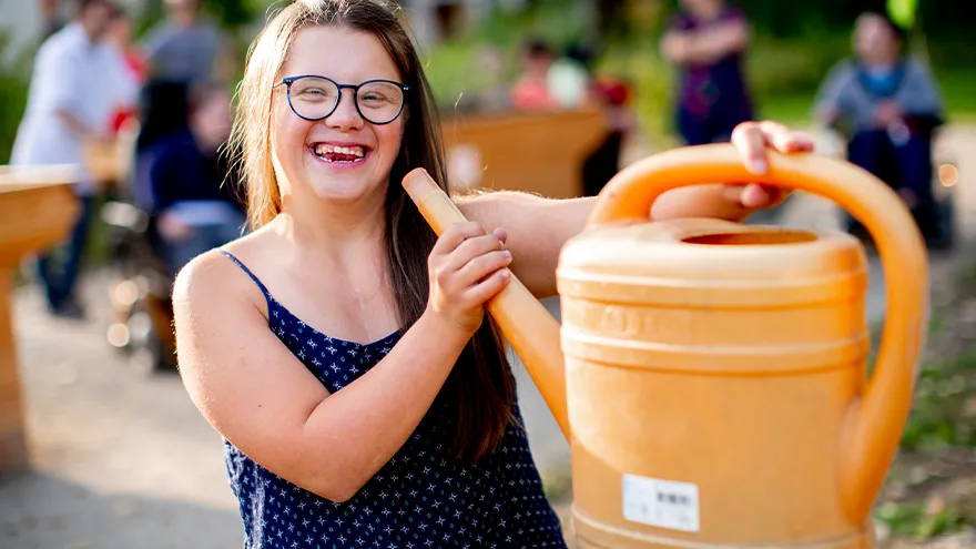 Eine lachende junge Frau mit Downsyndrom trägt eine Gießkanne