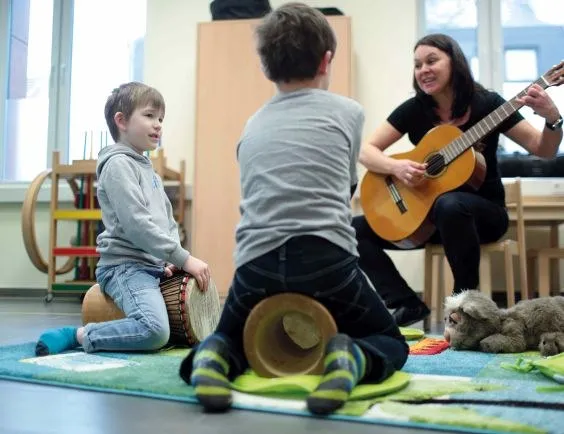 Eine Frau spielt Gitarre und singt, zwei Kleinkinder sehen zu.