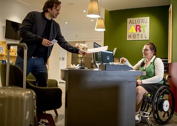 Ein junge Frau im Rollstuhl spricht mit einem Gast in einem Hotel.