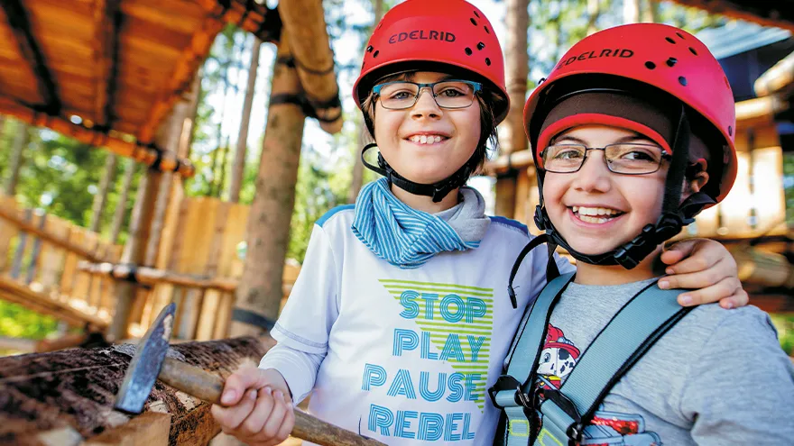 Zwei Kinder stehen in einem Abenteuerspielplatz im Wald, haben einen Helm auf dem Kopf, eines einen Hammer in der Hand.