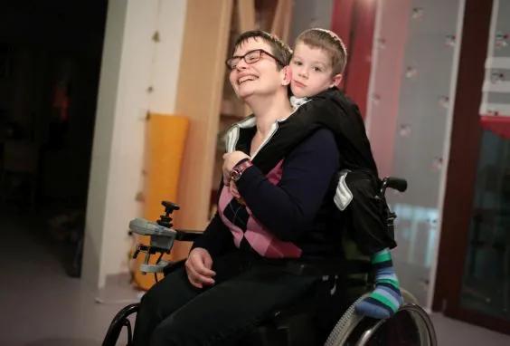 Eine Mutter im Rollstuhl mit ihrem Sohn. Das Kind steht hinten auf dem Rollstuhl.
