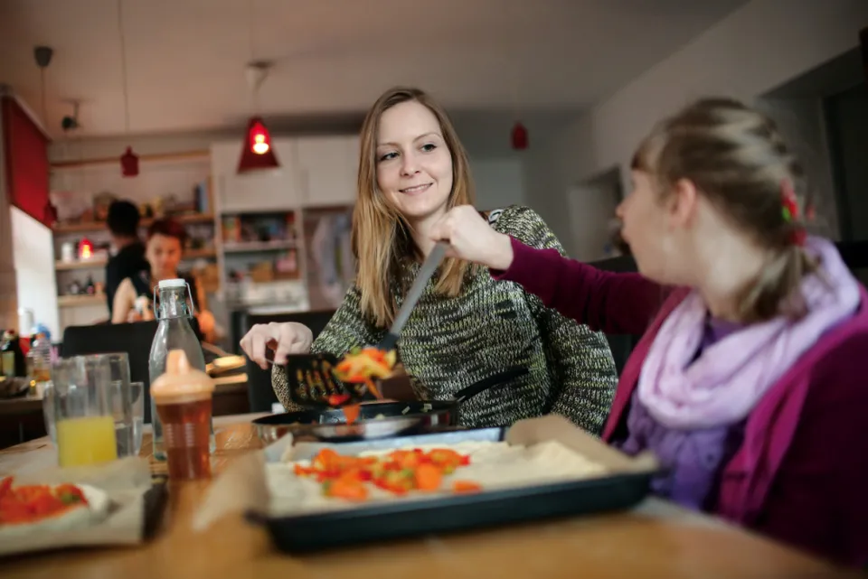 Eine junge Frau und eine andere junge Frau mit Behinderung belegen ein Pizza-Blech in einer Wohnküche.